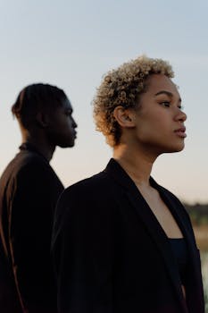 A fashionable man and woman pose outdoors in black suits during sunset, showcasing modern style.