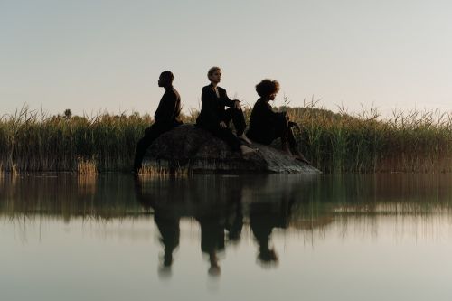 Free Three people elegantly posed on a rock by a serene lake with lush tall grass. Stock Photo