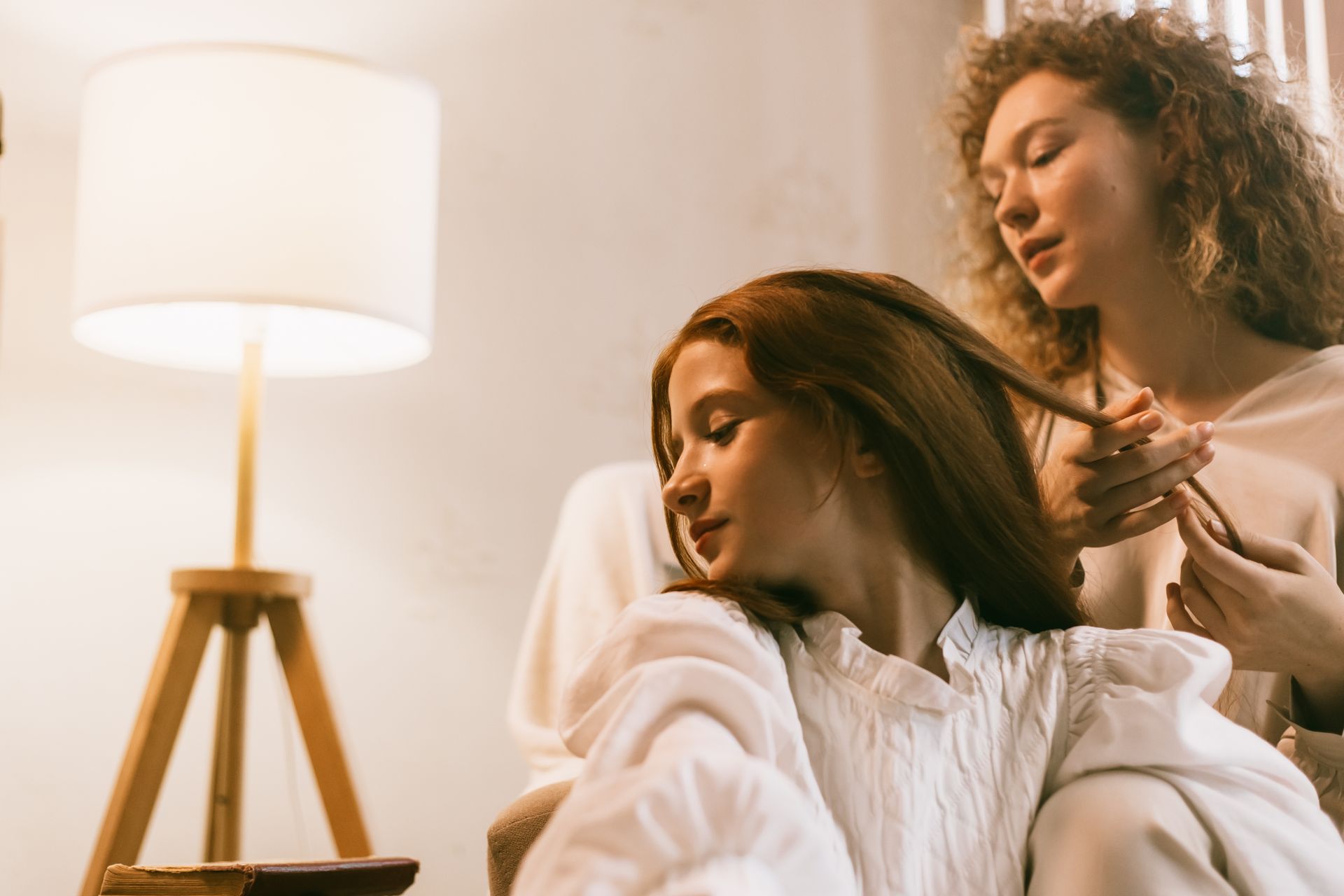 A woman braiding a girl's hair in a cozy living room with warm lighting.