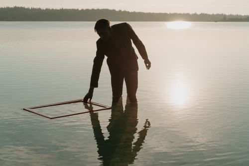 Free A man in a lake holding a frame at sunset, reflecting on the calm water surface. Stock Photo