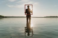 Man in Black Formal Attire Standing on Shallow Water while Holding a Red Wooden Frame