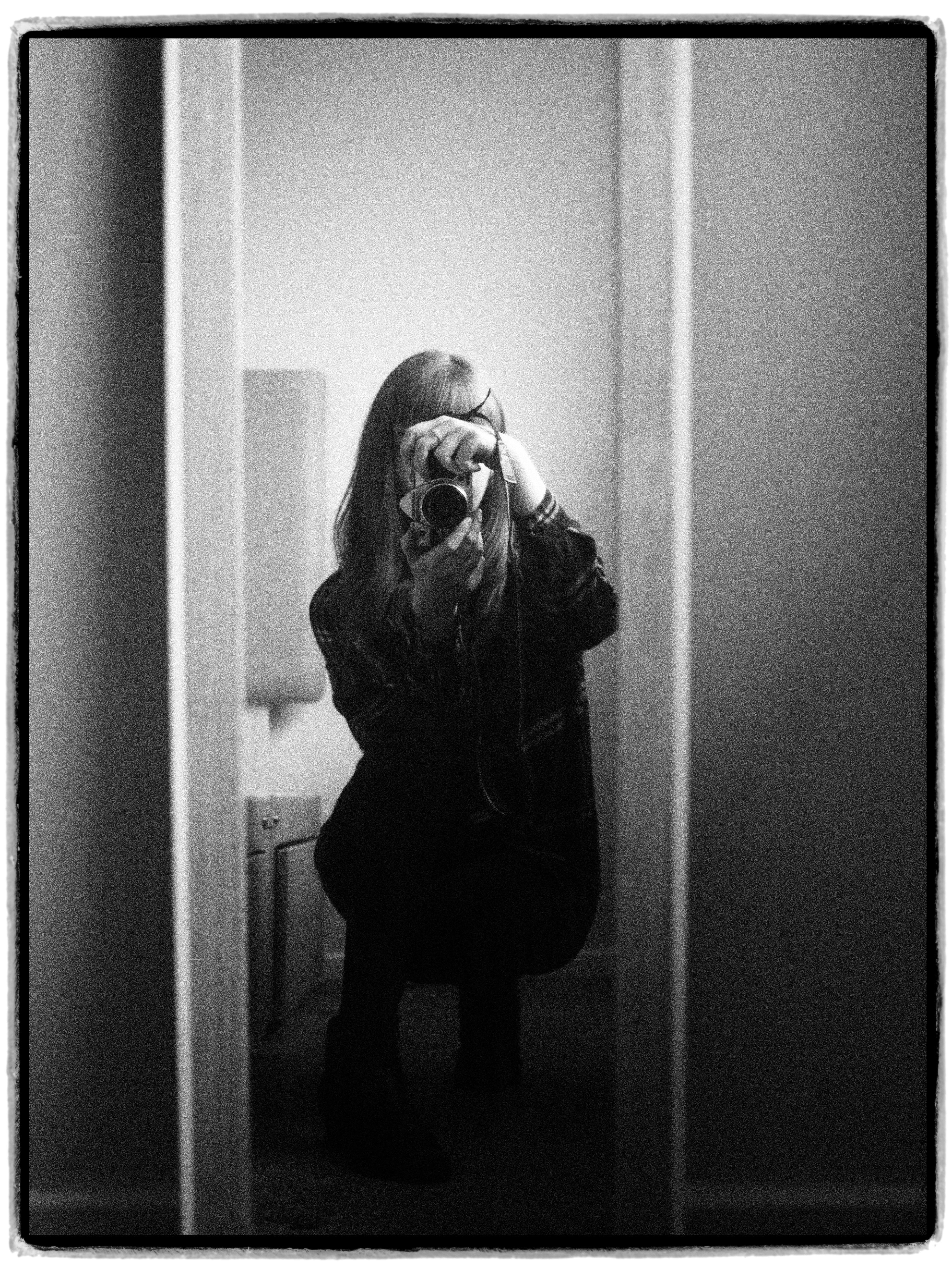 Black and white self-portrait of a woman crouching with a camera in front of a mirror.