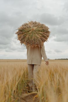 A person in a beige coat walks in a wheat field holding a grass umbrella, with a cloudy sky above.