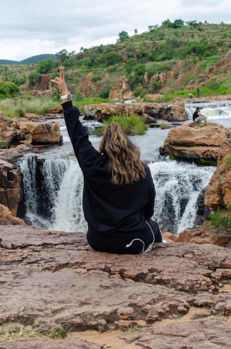 Back Of A Woman Sitting On Rocks Over A Waterfall