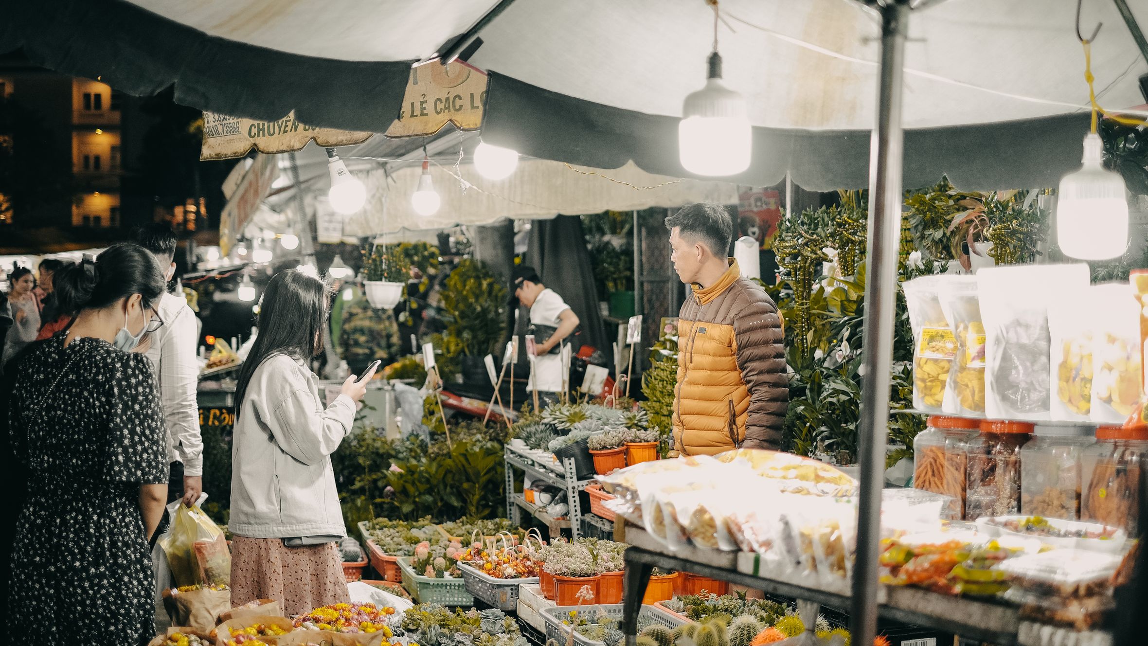 Lively street market scene with people shopping for fresh produce and goods under lights.