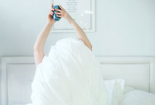 A person holding a coffee mug while relaxing in bed, covered with a white sheet.