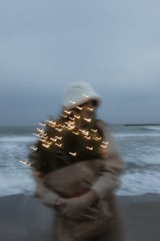 Romantic moody scene of a woman holding a lit Christmas tree on the beach.