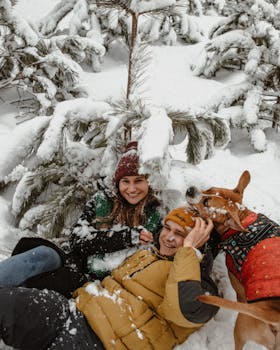 Happy couple laughing with a dog in winter snow, surrounded by fir trees.