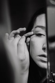 Black and white close-up of a person applying eyeliner in a mirror.