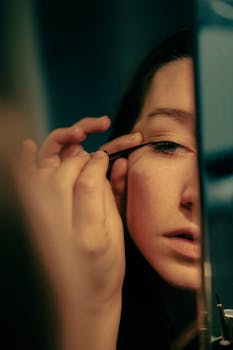 A young woman carefully applies eyeliner while looking into a mirror.