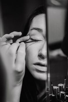 Black and white portrait of a woman applying liquid eyeliner in front of a mirror.