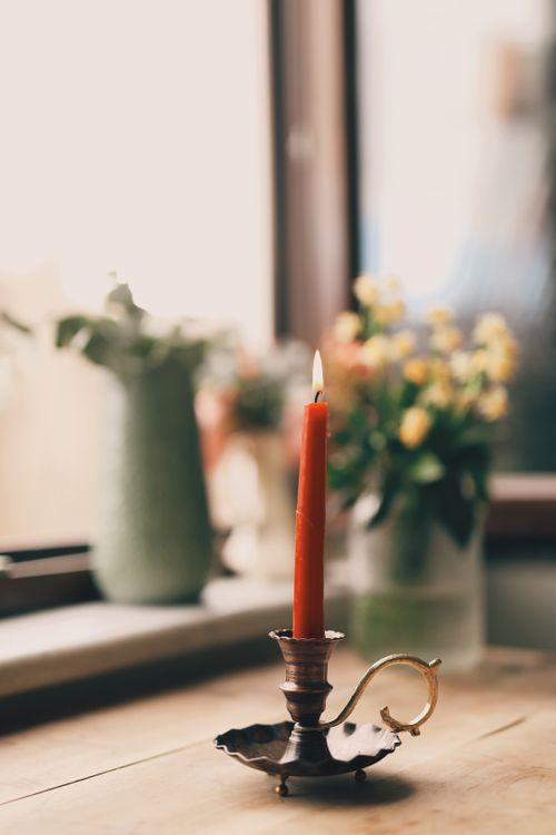 Free A vintage candleholder with a lit red candle on a wooden table by a window next to flowers. Stock Photo