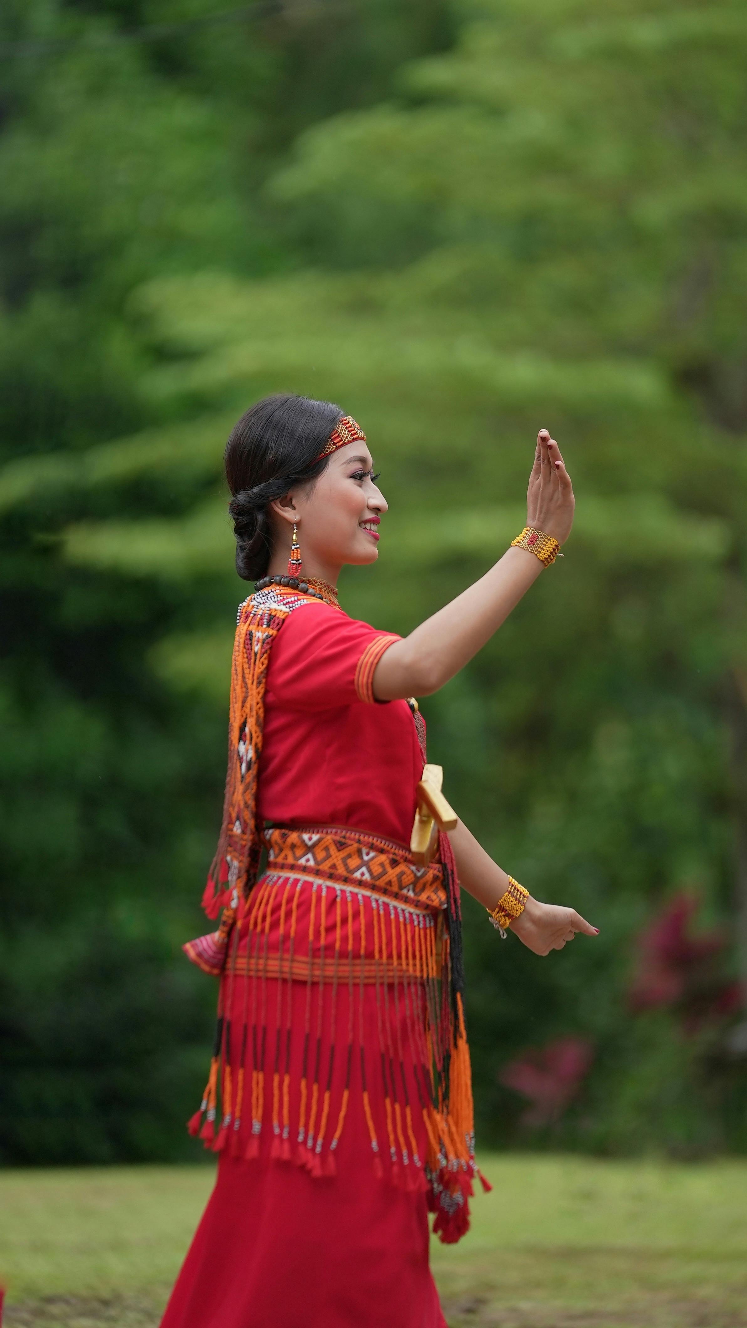 Portrait of a woman in traditional red attire, outdoors, smiling and waving.