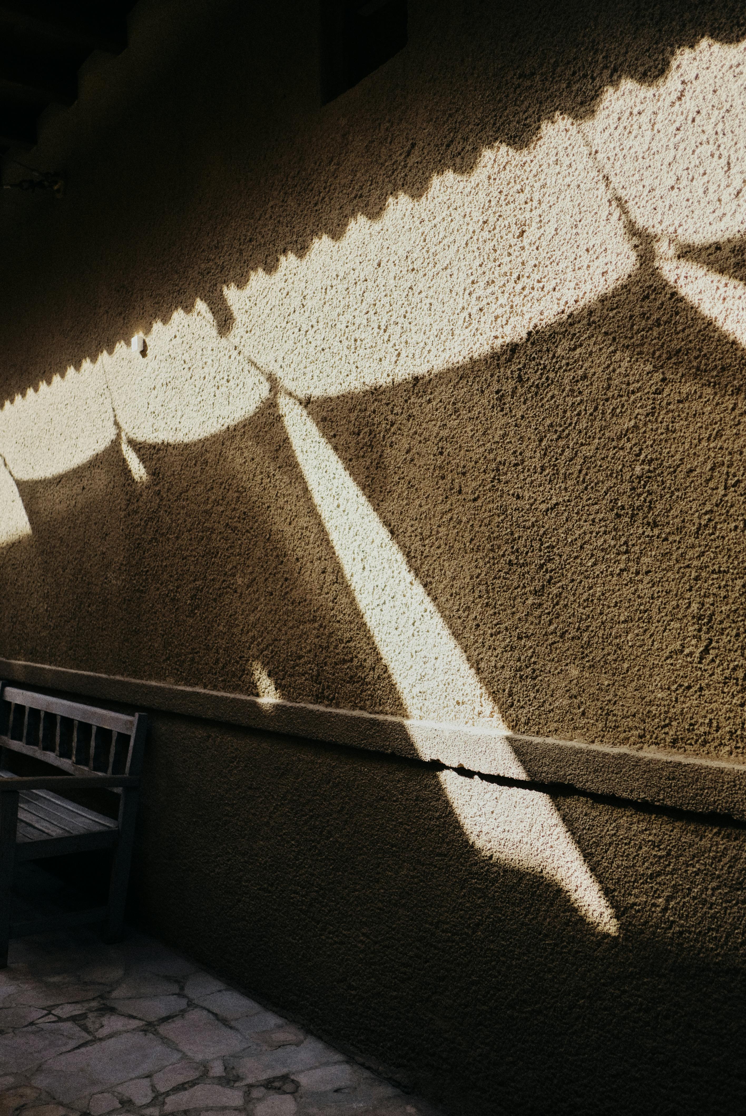 Free A wall with sunlit patterns creating shadows and a wooden bench outdoors. Stock Photo