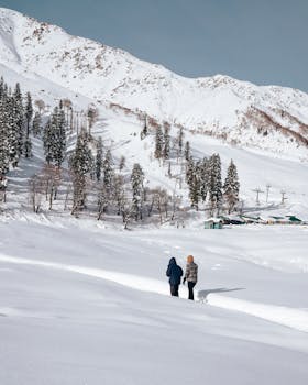 A couple walks through the snowy winter landscape of Gulmarg, surrounded by mountains and trees.