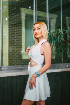 Stylish blonde woman poses in a white dress by a city street window, exuding modern fashion vibes.