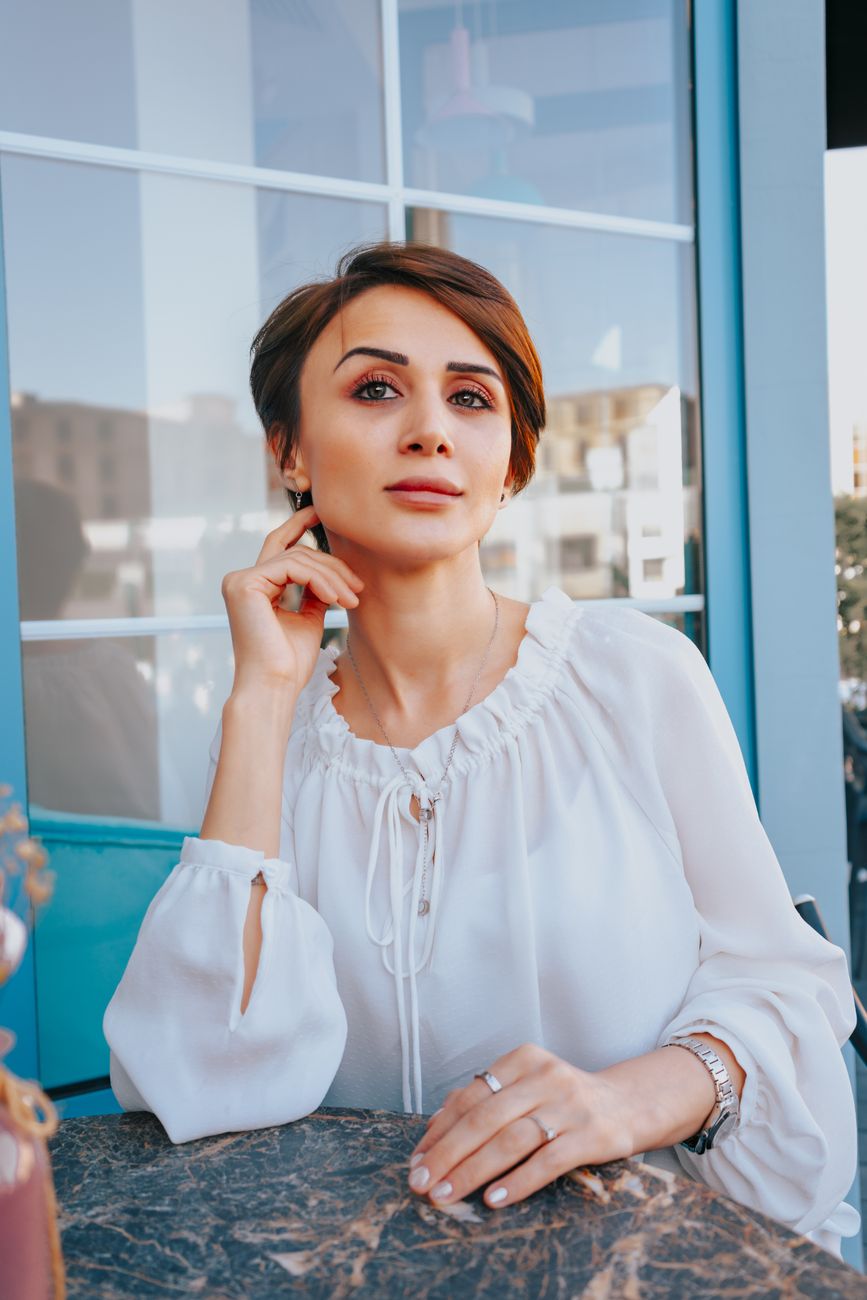 Portrait of a stylish woman in a white blouse sitting outdoors at a cafe table during the day.