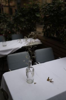 Minimalistic outdoor café table with glass bottle vase and delicate flowers, set against greenery.