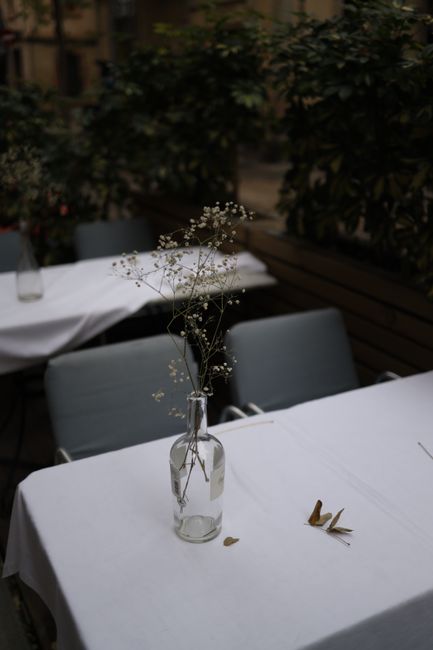 Minimalistic outdoor caf茅 table with glass bottle vase and delicate flowers, set against greenery.
