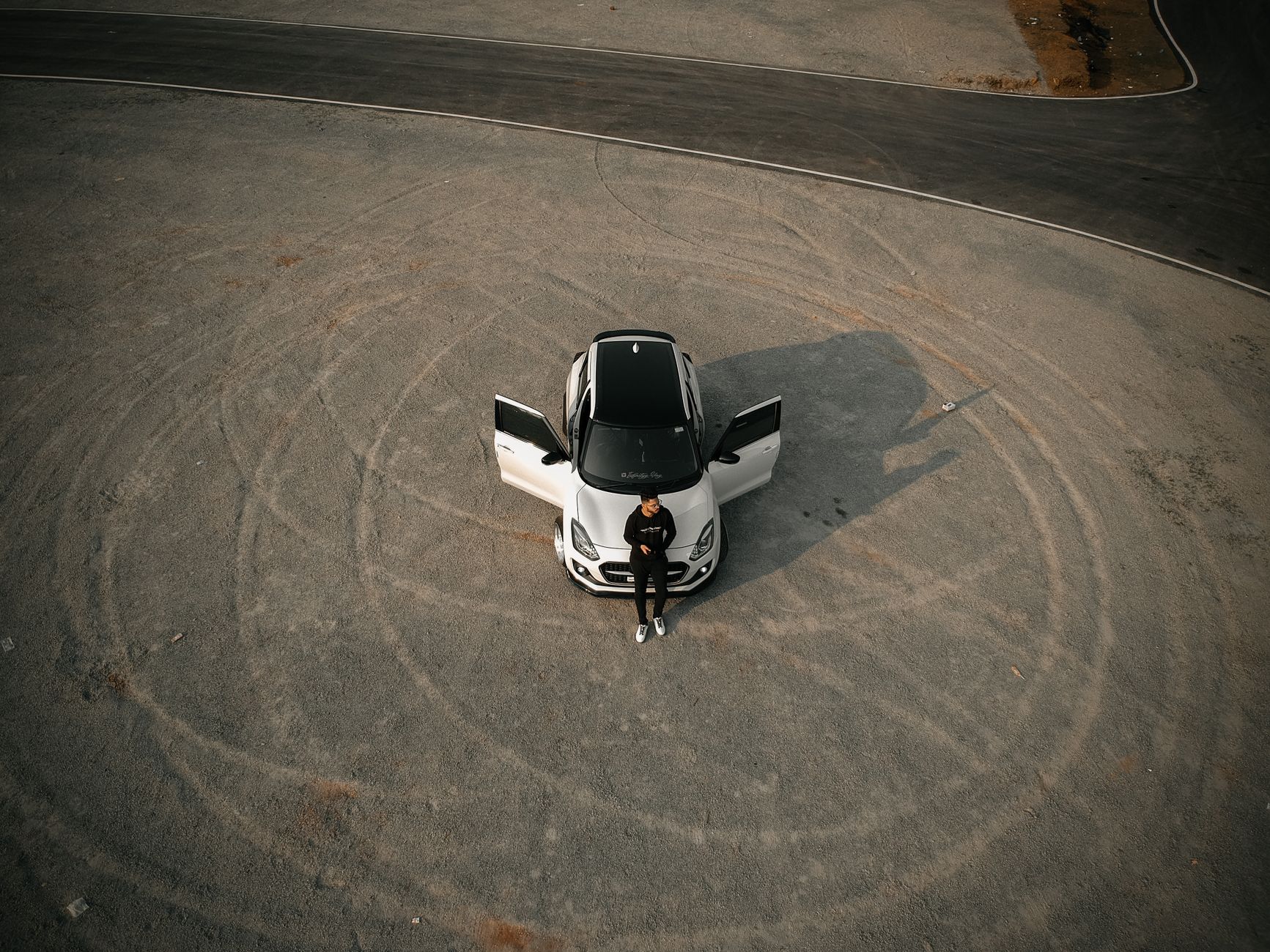 Aerial view of a man leaning on a white car with doors open, parked on a circular dirt track.