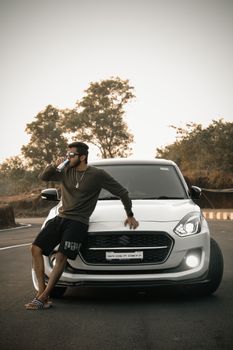 Man in casual attire drinks next to a parked white car on a roadside.