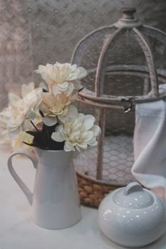 Delicate white flowers in a ceramic pitcher beside a decorative birdcage, creating an elegant still life scene.