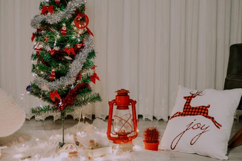 Free Cozy Christmas tree with decorations and a red lantern beside a joy-themed pillow indoors. Stock Photo