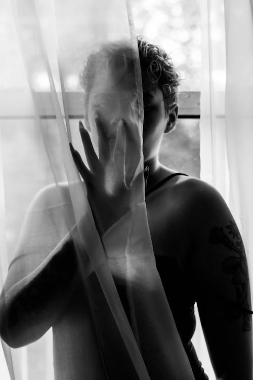 Grayscale Photo of a Woman Hiding Behind the Curtain