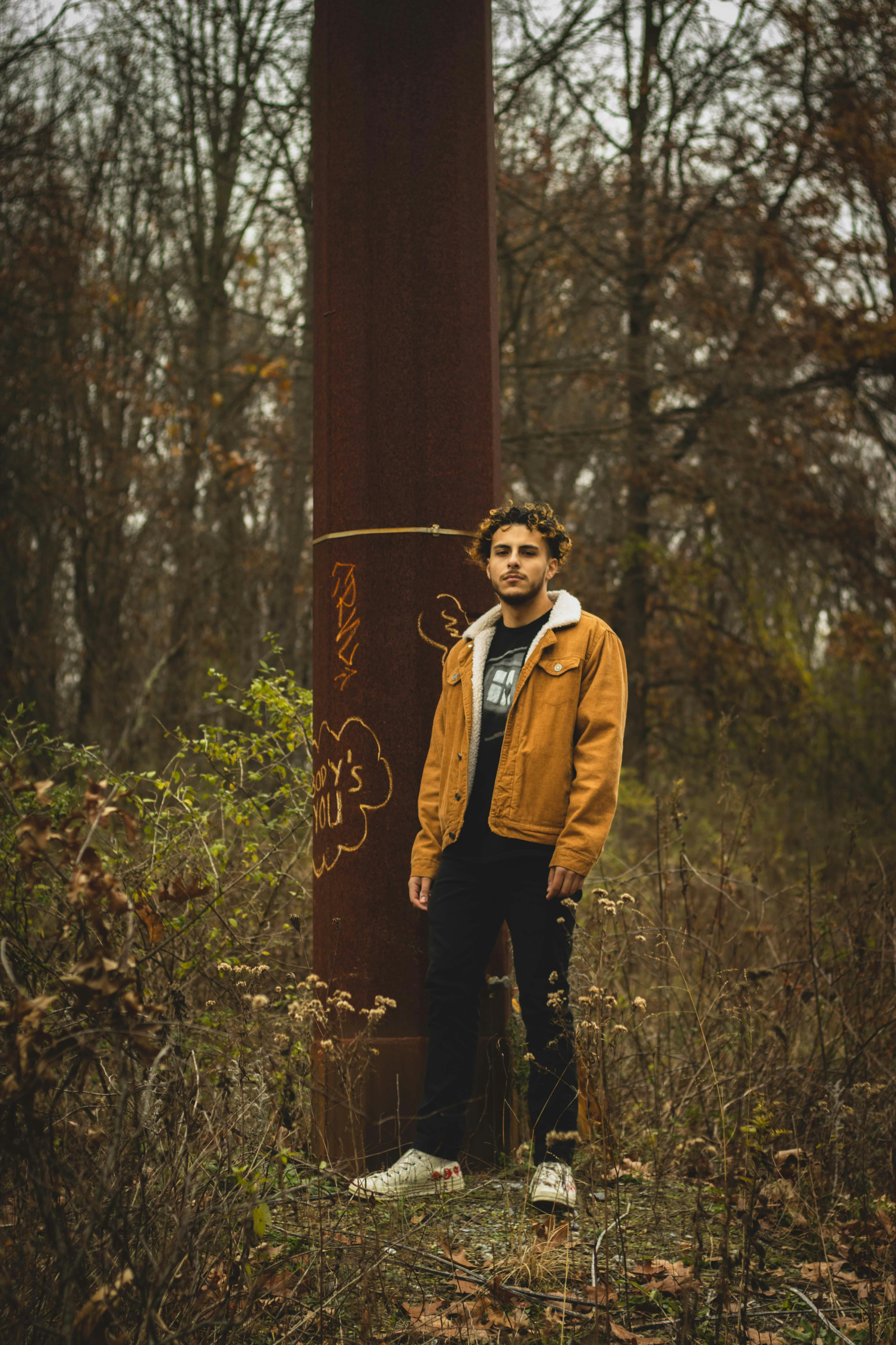 A young man with curly hair stands near a rusty pole in a wooded area during autumn.