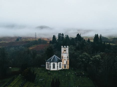 Drone shot of a historic church surrounded by misty hills and dense forest in Scotland.