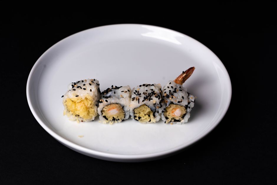 Sliced shrimp tempura sushi rolls arranged on a black plate, showing the crispy fried shrimp tail, avocado, and cucumber filling, drizzled with spicy mayo and eel sauce and topped with sesame seeds.