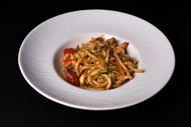 Close-up of stir-fried udon noodles with vegetables on a white plate against a dark background.