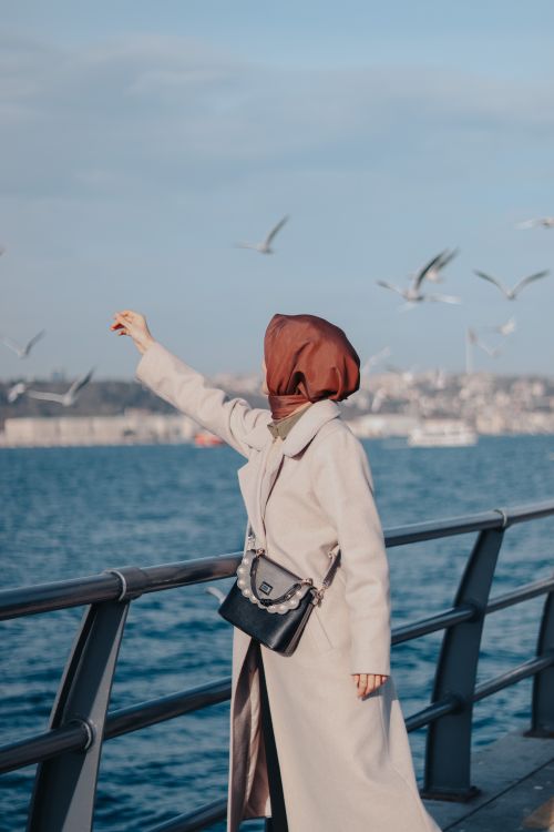 Free A woman in a beige coat feeds seagulls on a waterfront promenade, offering a serene coastal scene. Stock Photo