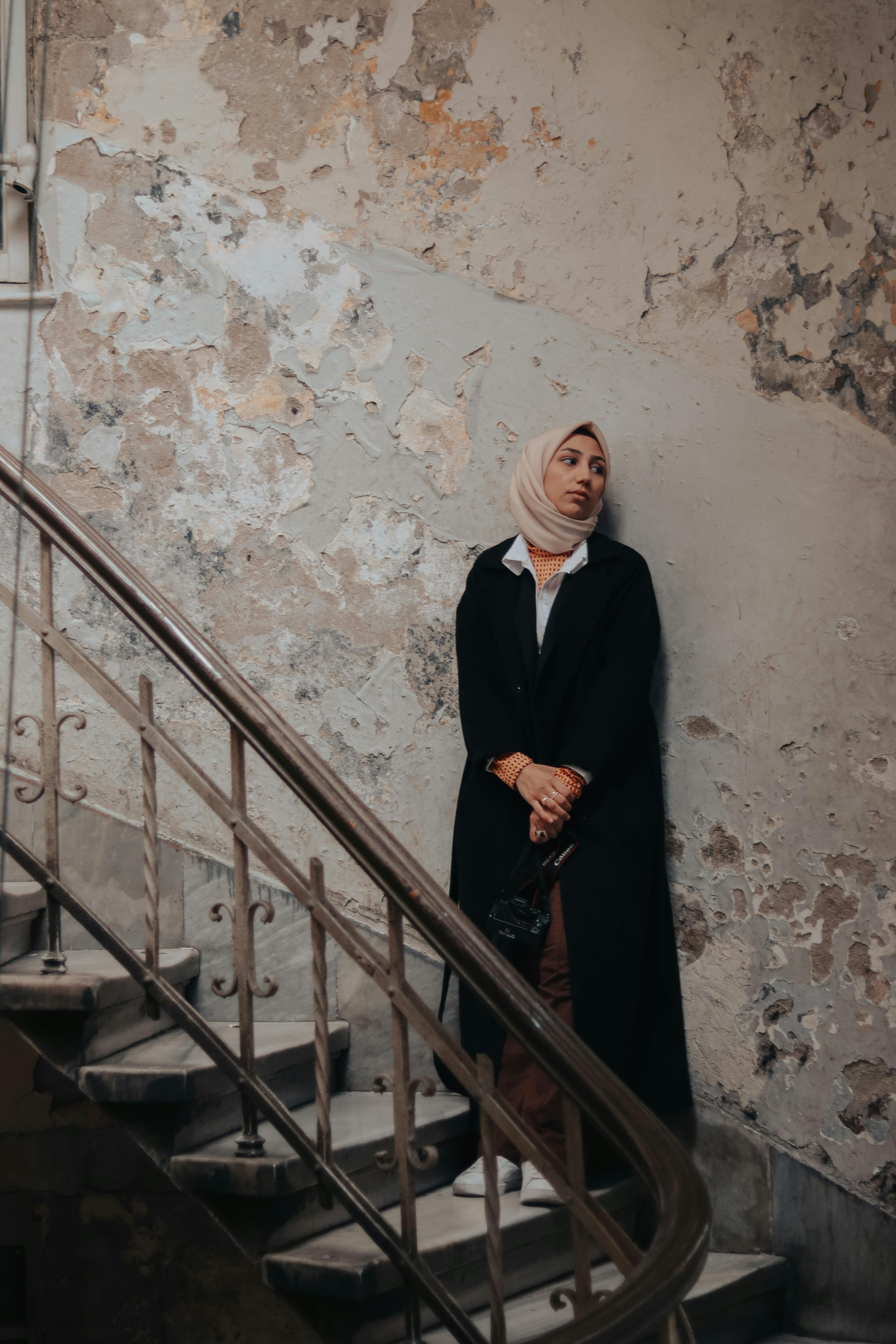 A woman in a headscarf leans against an aged wall by a staircase, exuding elegance and grace.