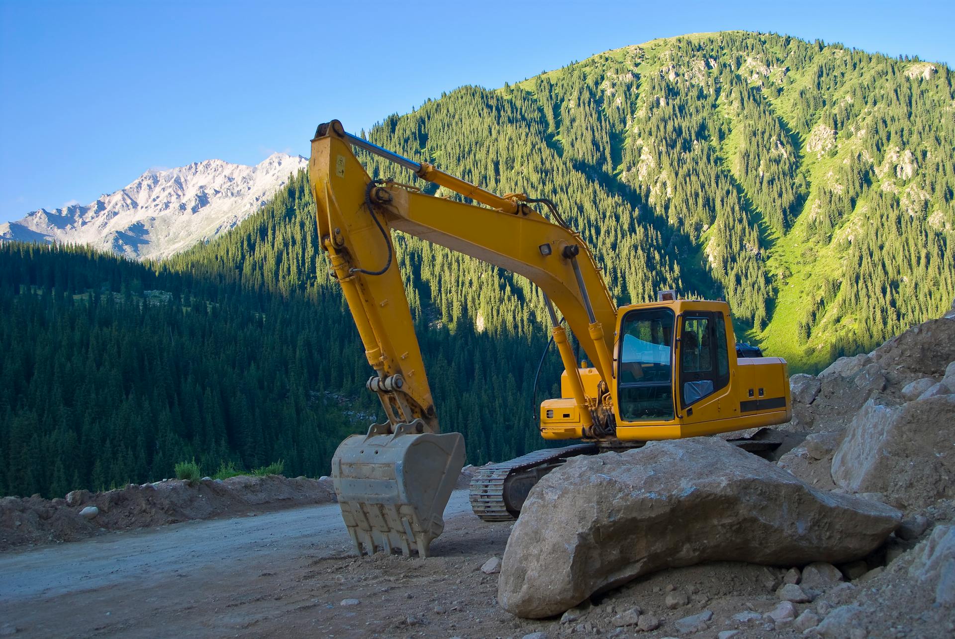 Yellow excavator on a rocky dirt road with forested mountains in the background, a scenic outdoor construction setting.