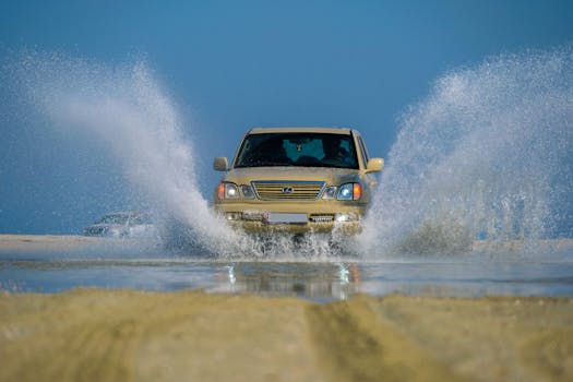 A beige SUV splashes through a puddle on sandy terrain under a clear blue sky.