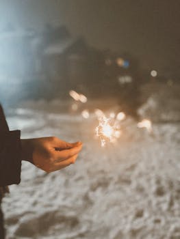 A person holding a sparkler in a snowy landscape at night, creating a festive atmosphere.