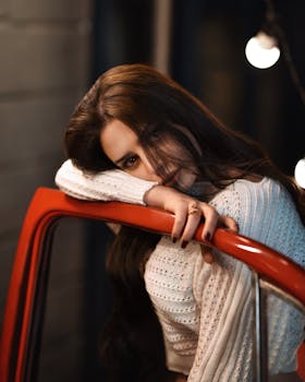 A woman in a white sweater leans on a car door, exuding elegance and mystery in warm lighting.