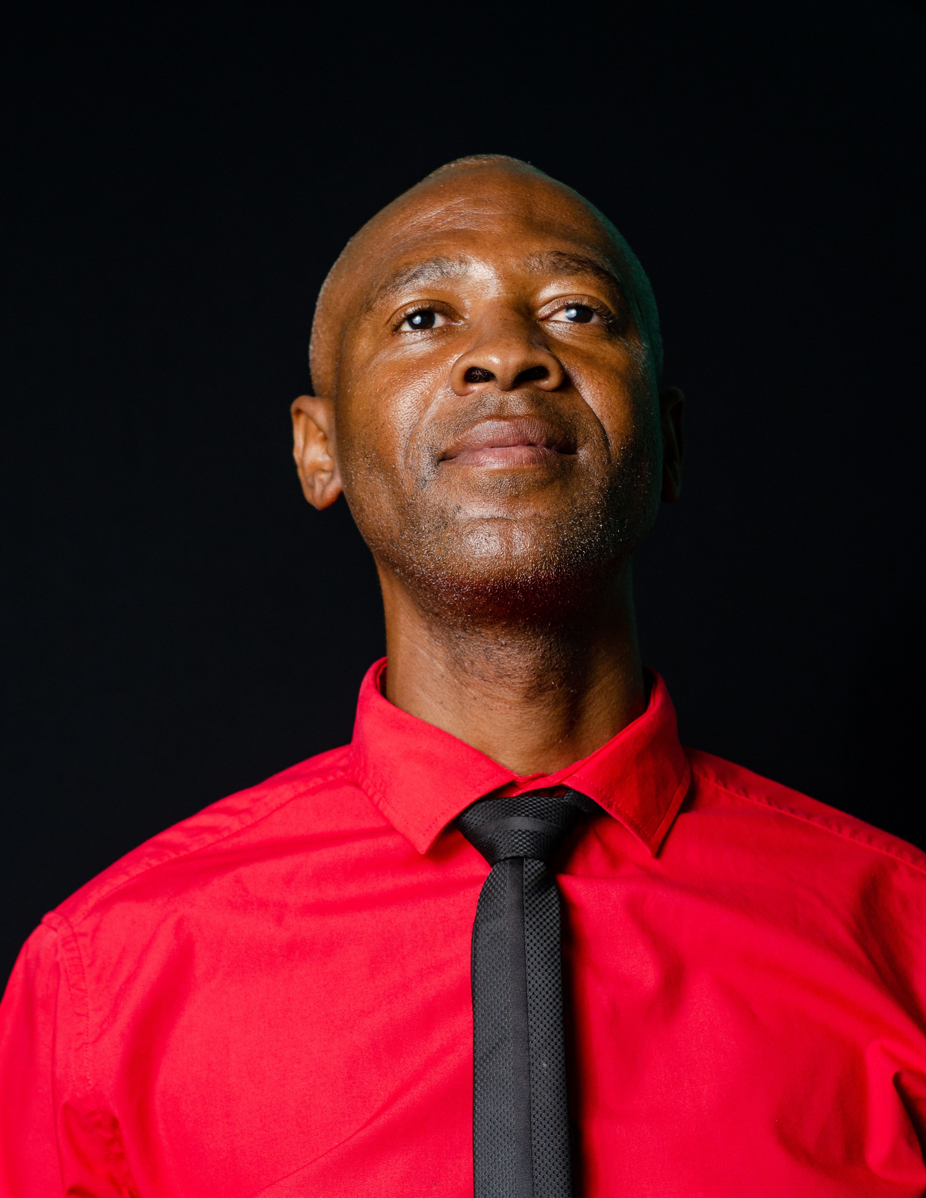 Portrait of a confident bald man wearing a red shirt and black tie against a dark background.
