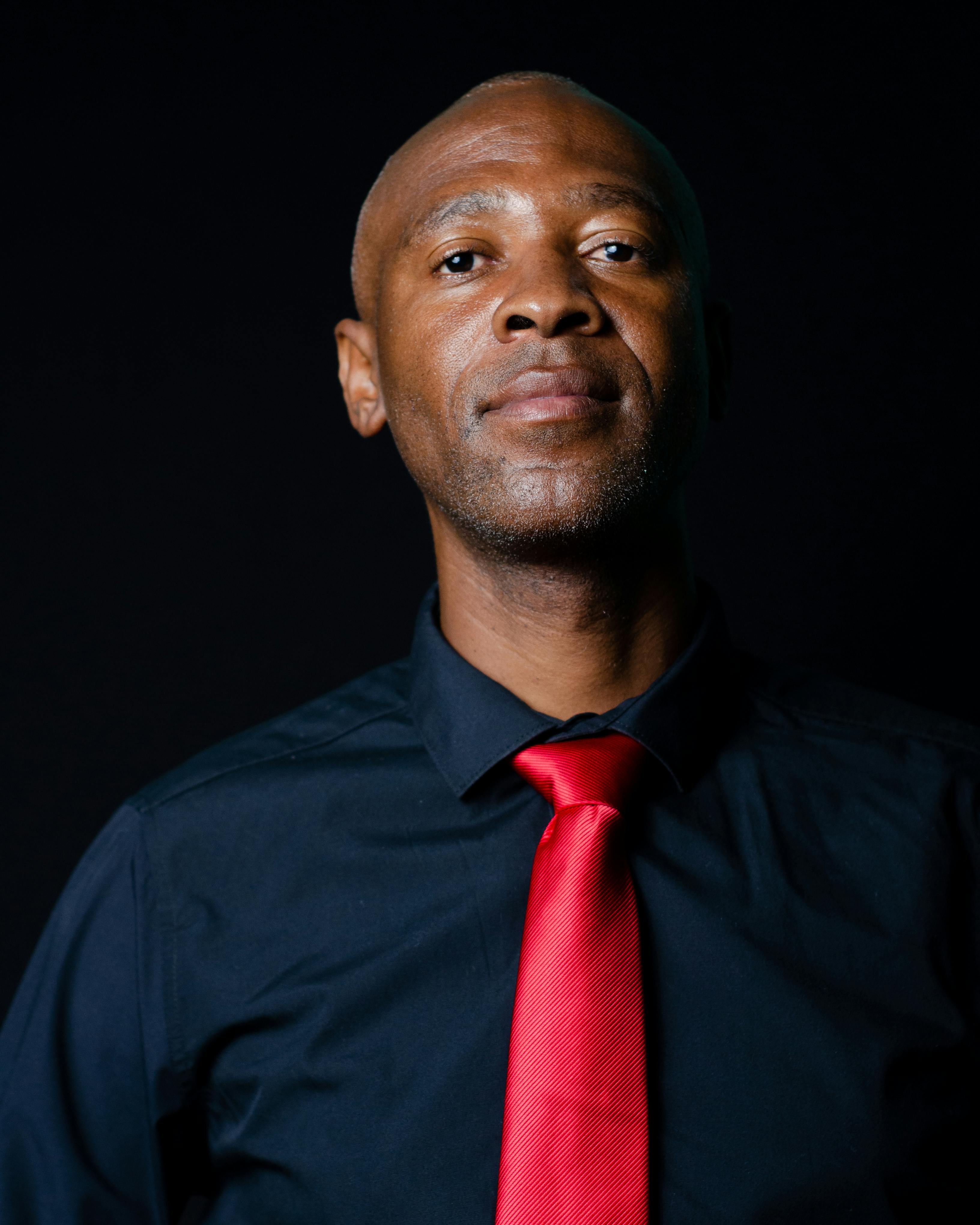 Free Portrait of a bald African American man wearing a red necktie and black shirt, set against a dark background. Stock Photo