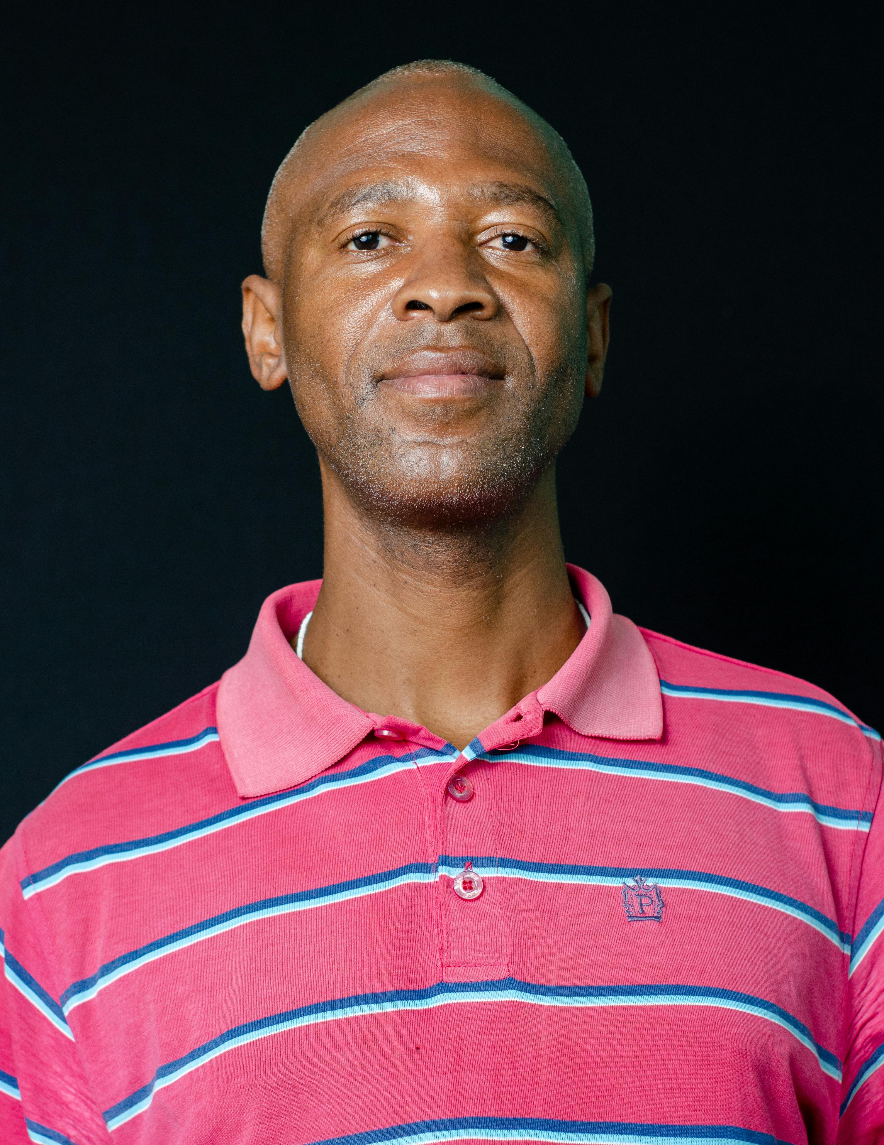 Close-up portrait of an African American man wearing a pink striped polo against a black background.