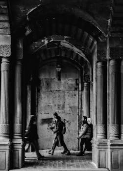 Black and white photo of people walking under a gothic archway in a city setting.