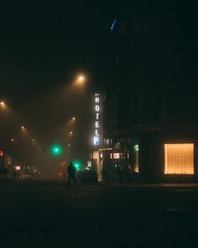 A foggy night street scene featuring a glowing hotel sign and street lights creating a cinematic ambiance.