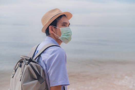 Man wearing a surgical mask and fedora backpacking on a serene beach, embracing the new normal.
