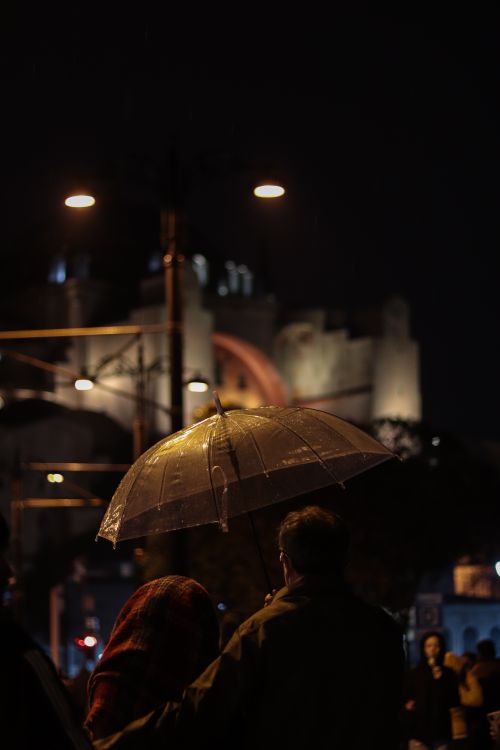 Free Couple walks under an umbrella on a rainy city night, streetlights casting a warm glow. Stock Photo