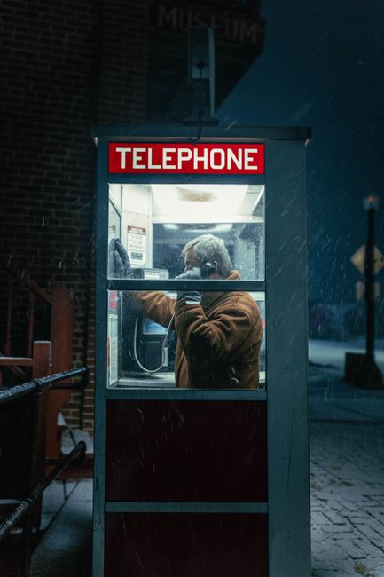 A man talking on the phone in a classic red telephone booth during a snowy night.