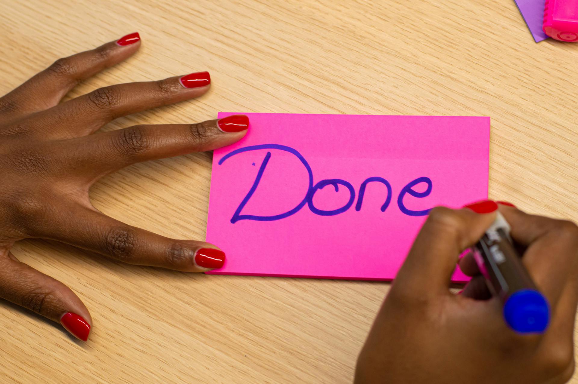 Close-up of a hand writing 'Done' on a pink sticky note with red nail polish.