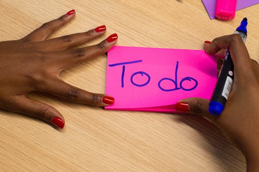 Hands writing a 'To do' note on a pink sticky note with red manicured nails.
