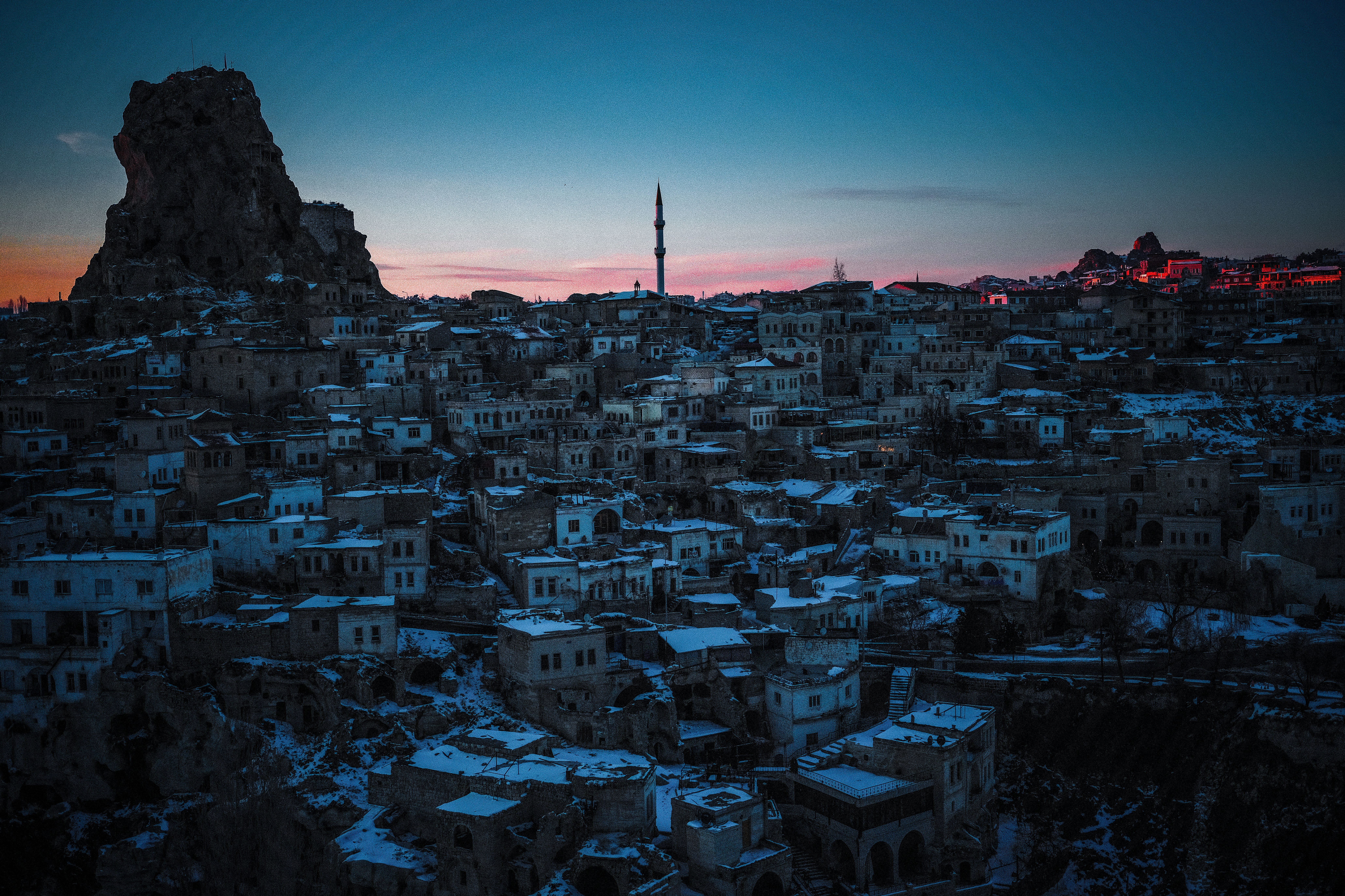 Aerial view of Cappadocia's ancient town under snow with towering rock formation at twilight.