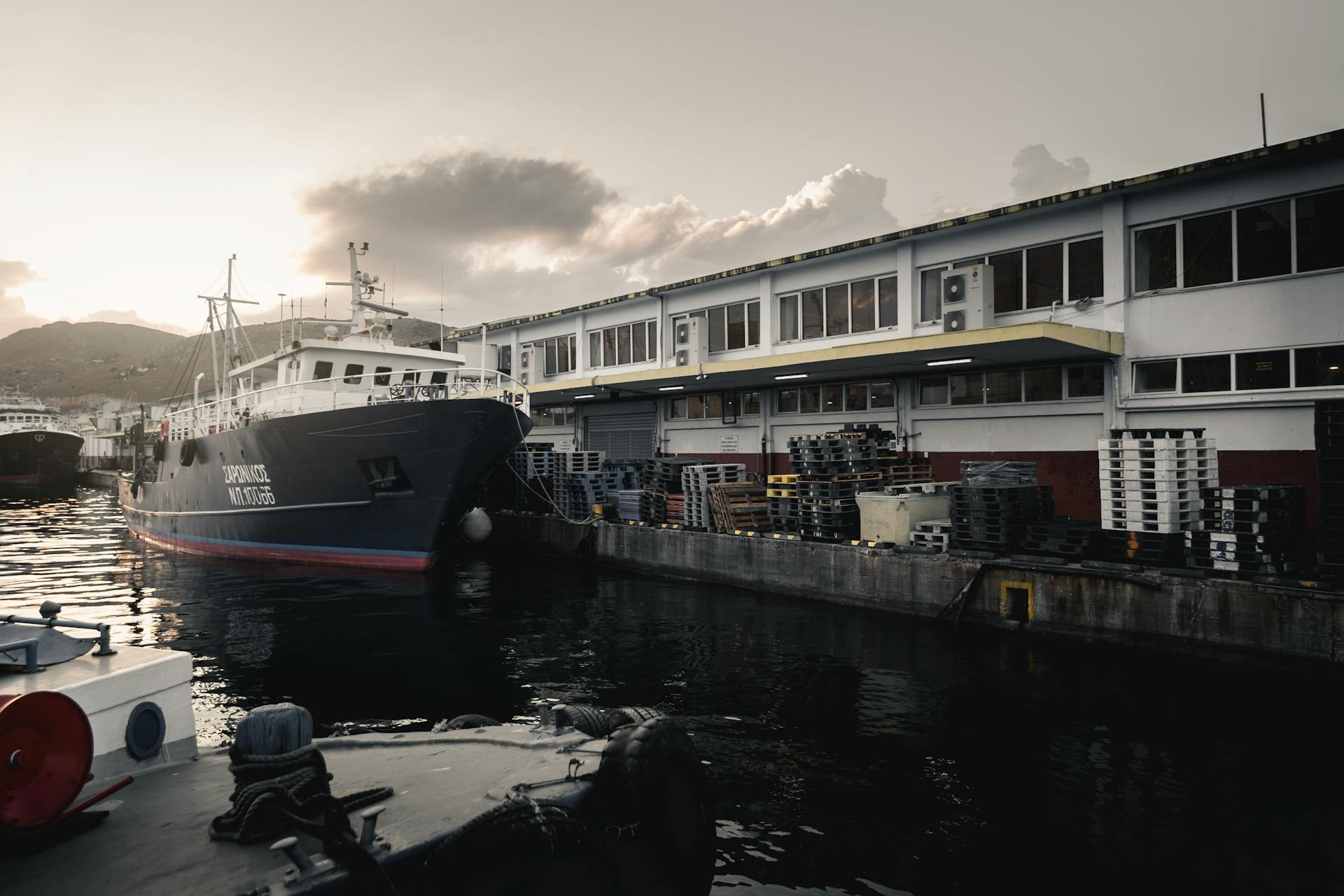 Cargo ship docked at an industrial port surrounded by pallets at sunset, highlighting maritime activities.
